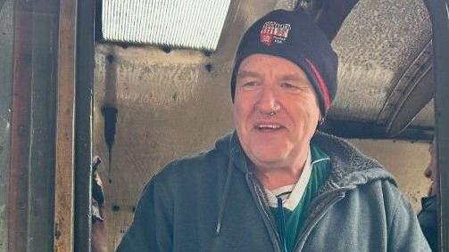 Martin Sullivan: a smiling, wearing a dark hat, dark hoodie and top, and standing in the doorway of an old train carriage.
