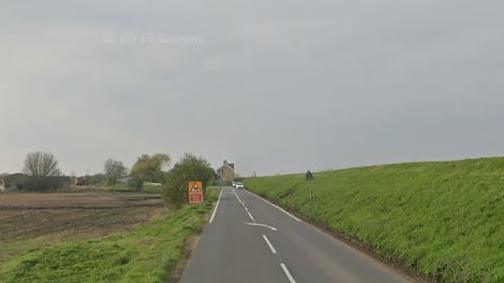 Branch Bank, a fen road with a high grass covered bank to the right and low-lying arable fields to the left. The road is single carriageway. In the distance is a house and a white car is approaching. 