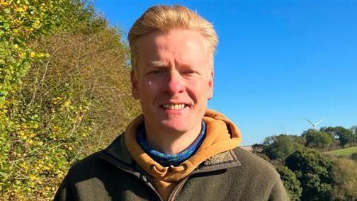 Matthew Snedker, who has blonde hair and is wearing a yellow hoody and green coat, stands in the foreground to the left, with a field in the background and a clear blue sky overhead.