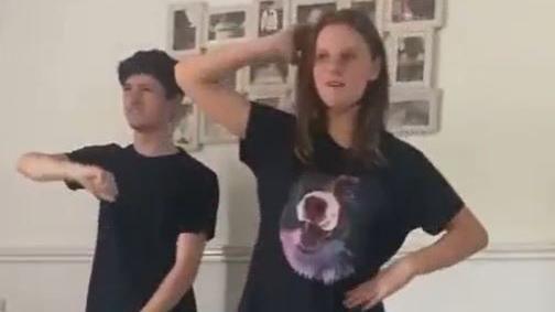 Still taken from a video of a boy and a girl pulling dance moves in a living room. She is wearing a black t shirt with a picture of a dog on it, he is in just a plain black t shirt.