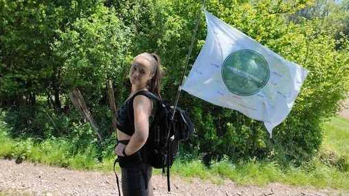 Kit Birks is standing smiling side-on to the camera, she has brown hair tied back into a ponytail, and is wearing black sport shorts and bra. On her back she is carrying a black pack-back with a flag adorned with the names of suicide victims.