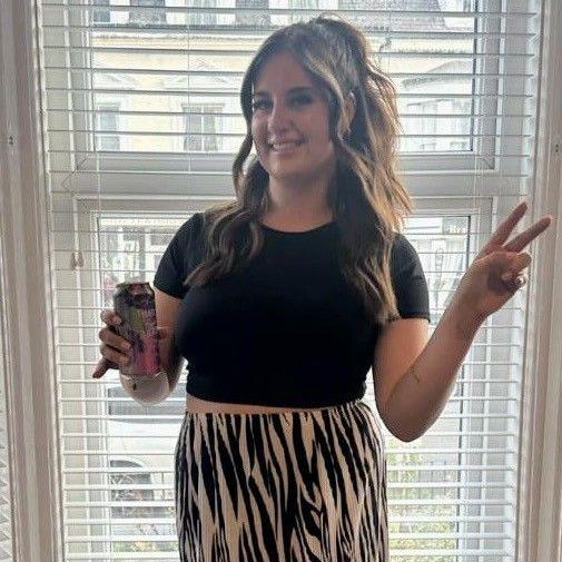 Paige Roberts standing in bay window wearing zebra print trousers and a black belly top. She is smiling and holding a can in one hand and throwing up the peace sign with her other 