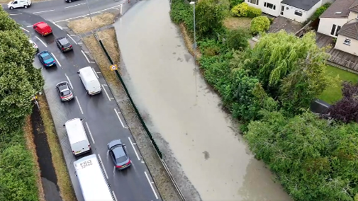A view of a road approaching a roundabout with traffic on the left handside. The right hand side of the road is covered in water. 