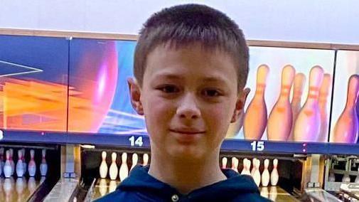 Leo looking straight at the camera. Ten-pin bowling is behind him with an alley leading to a set of pins.