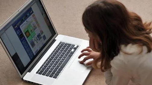 A young girl whose face cannot be seen lies on the floor as she looks at a laptop