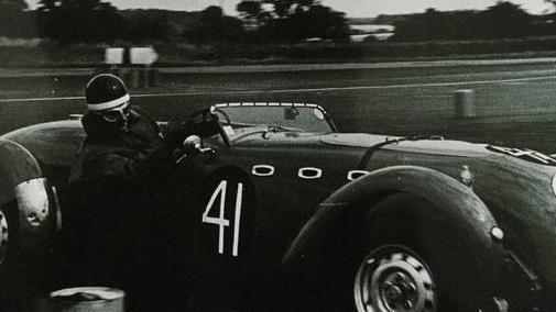 The picture shows a vintage racing car on a track, captured in black and white. The car is an open-top sports racer with a sleek, aerodynamic body and prominent front wheel arches. It has the number 41 painted on the side. The driver is wearing protective gear, including a helmet and goggles, and is gripping the steering wheel while leaning into a turn.
