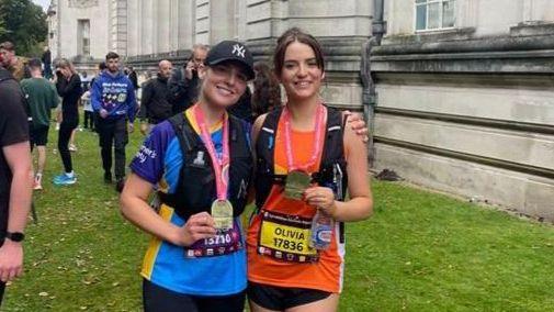 Olivia Browne and her friend Grace Davies stand in front of Cardiff's National Museum. They are holding medals. Olivia is wearing a pink shirt and a black running vest. Grace is wearing a blue shirt and a black running vest. Both have half marathon labels with their name and number on.