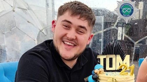 A man with short brown hair and a black shirt smiling while sitting on a blue chair and next to a cake that has "Tom" and "21" on top of it.