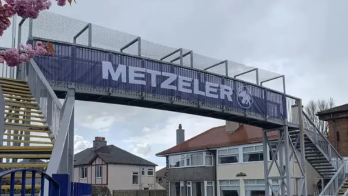 A metal footbridge  over a road. It has steps leading up to it on both sides and is roofed over with mesh. There is a blue advertising banner pulled across it and houses and pink cherry blossom can be seen at wither side.