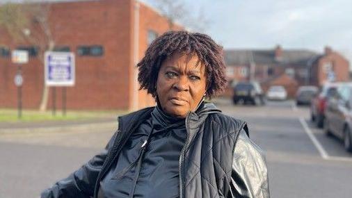 A woman is standing in a car park looking to her left. She is wearing a black padded jacked with a black sports top underneath. 