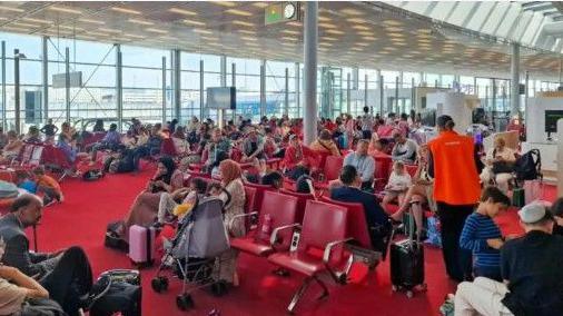 Scores of passengers can be seen sitting on rows of red seats at the gate. They are next to a very large window.