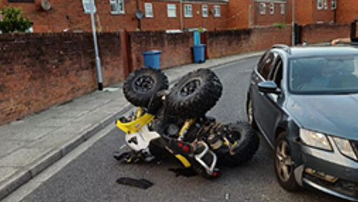 Yellow and white quad bike flipped in the road in Liverpool next to a silver car. Behind the quad bike is a brick wall with blue wheelie bins out and houses behind the wall.