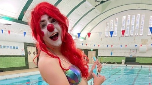 A woman is dressed in a purple, shining mermaid costume. She is wearing a red, long wig and her face is made up to look like a clown, with a red nose. She is standing by an indoor pool and is laughing into the camera.