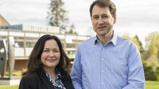 A photo of Tanya Ednan-Laperouse, a woman with brown hair wearing a black jacket and blue and white shirt, stood next to her husband Nadim Ednan-Laperouse, who is wearing a blue shirt. There is a building and greenery behind them.