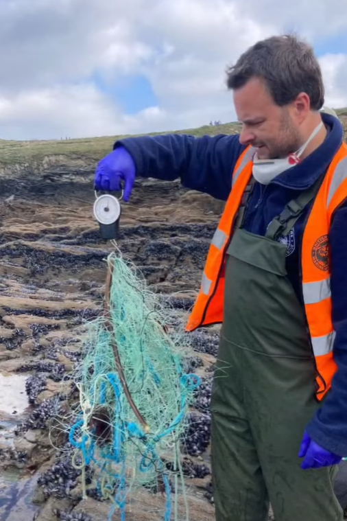 Dan Jarvis is standing on the beach holding the mass of fishing net up on a scale. It looks like an armful of net. He is wearing gloves, a high-vis waistcoat and a mask is hanging around his neck. 