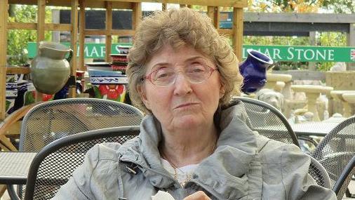 Pat sitting outside at a garden centre cafe wearing a green coat 