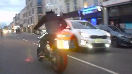 Motorcyclist riding bike in CCTV image taken from behind. He is wearing a white helmet and dark clothing with a white reflective line going down his trousers.