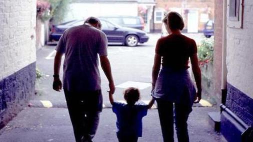 Library shot of two parents holding the hands of a child (seen from the back) heading towards a street