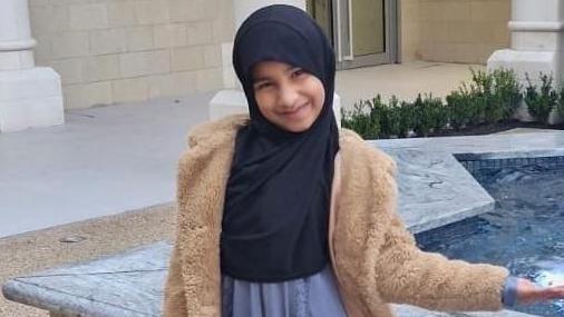 Inaayah Makda, wearing a brown furry jacket and a headscarf, smiles at the camera as she stands next to a water fountain in the shape of a star.