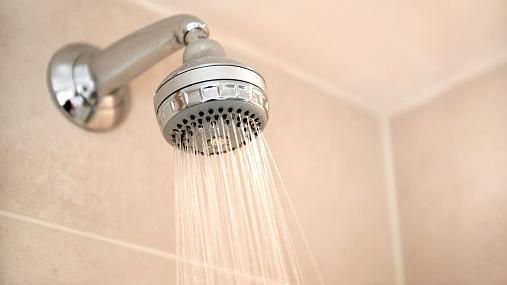 Silver shower head with water coming out in cream tiled bathroom.