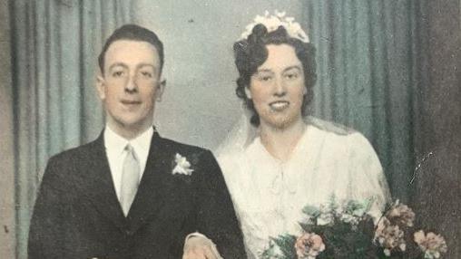 Image taken of Tessa Gay and her husband in their wedding outfits at a photograpy studio. He is on the left wearing a smart black suit and grey tie while she is in a white dress and wearing a floral headband and wedding veil behind her, holding a big bouquet of flowers.