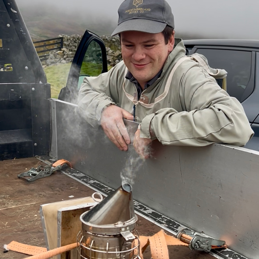 Luke Hutchinson is leaning over the back of a open-back truck and smiling. He is looking at a metal bee smoker.