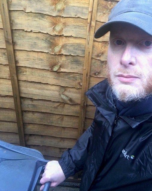 A man with a ginger and white beard, wearing a pale blue hat and a dark jacket, stands next to a fence while holding a wheelie bin.