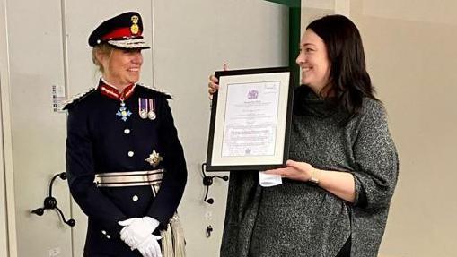 King's representative Dame Hilary Chapman, the Lord-Lieutenant for South Yorkshire, wearing a navy uniform with medals, military cap and white gloves, presents the framed award certificate to the Sunday Centre's chair Kelly Lingard wearing a grey top