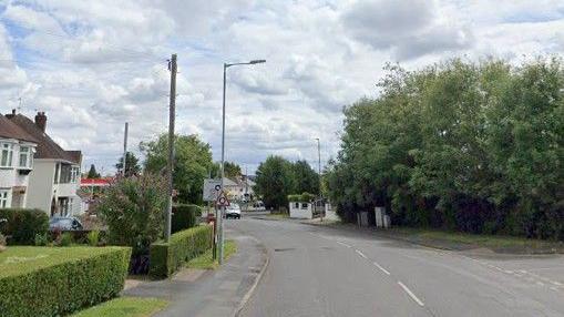A street, viewed on the approach to a roundabout. There aer houses either side, with low walls or hedges on the boundary of their driveways.
