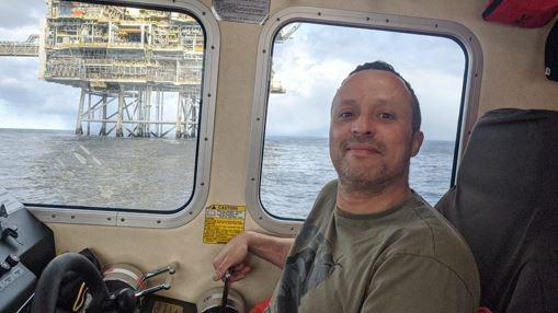 A man with receding hair, a khaki green t-shirt, sitting in a boat, smiling at the camera, with an oil rig at sea behind him