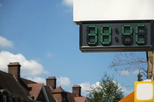 A digital thermometer in London showing the temperatures during the UK heat wave. It reads 38 °C. In the background there are trees and houses, but only the roofs and chimneys are visible.