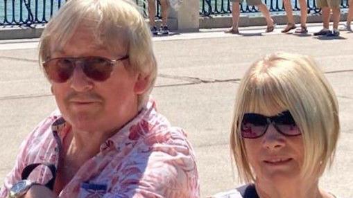 A couple are smiling at the camera. It looks like a sunny day at the sea side