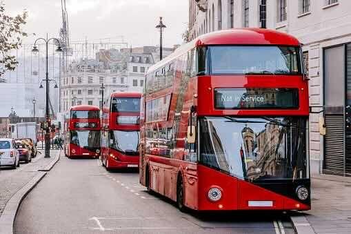 Three red Transport for London buses on a street in central London. The bus at the front is out of service and parked in a bus lane. In the background there are lampposts, buildings and parked cars on the opposite side of the street.