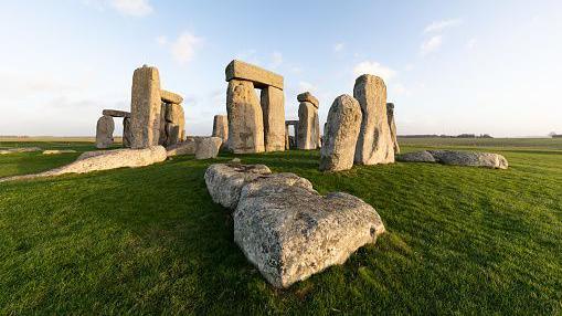 The Stonehenge circle of stones in the early morning light.  There is lots of green grass and there are shadows across some of the stones.