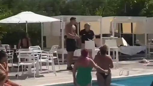 Kian Harrett stands topless in black shorts looking on after throwing a chair at a person by a swimming pool.