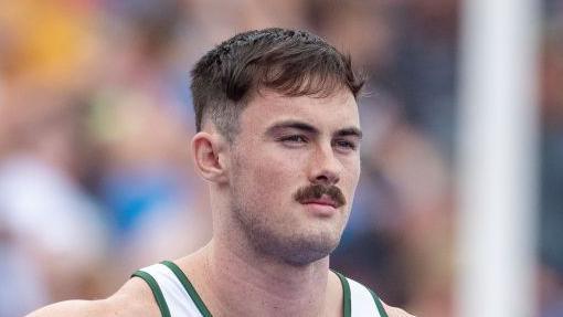 A close-up of Joe Chadwick's head and shoulders as he stands waiting on a start line. He has short dark hair and moustache and wears a white running vest with green piping.