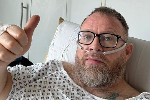 Neil Maggs is in a hospital bed wearing patient clothing. He has a tube attached to his nose and is giving a thumbs up. 