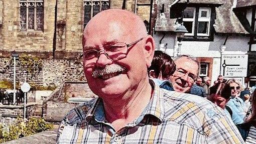 A bald headed man in a checked shirt with glasses and a moustache stands on a bridge on a sunny day