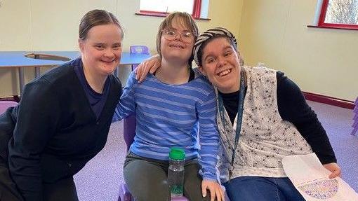 Three women sitting down hugging each other with bright smiles in a meeting room. 