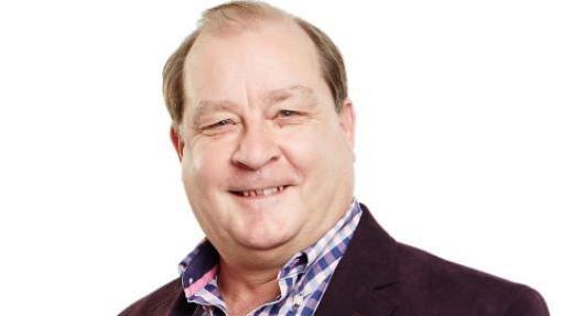 Andy Ball with brown hair smiling wearinga pink and blue checked shirt and dark brown blazer