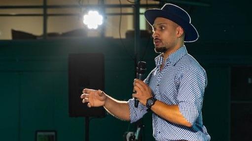 Raghoo is speaking to students at the event, holding a microphone. He is wearing a blue and white checked shirt and a hat. 