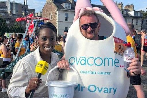 Craig McMurrough in his Mr Ovary costume holds a fundraising bucket in one hand and a bottle of water in another. A woman holding a yellow BBC Sport microphone stands to his right and is smiling at the camera.
