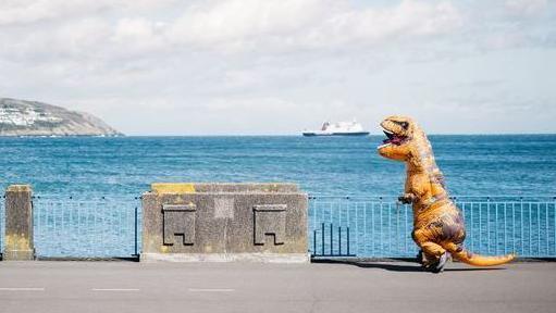 A runner dressed in an inflatable dinosaur costume makes their way along Douglas Promenade. There are stone blocks between metal railings and the the blue sea can be seen in the background with a ferry sailing by.