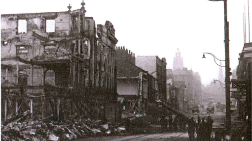 A black and white picture shows a street filled with bombed buildings and rubble. Several people appear to be standing on the right, huddled in a small group.