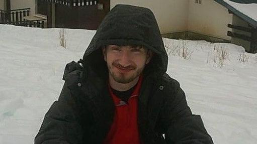 Matthew Pring poses for a photo in an outdoor area covered with deep snow. He is squatting and smiling and wears a dark anorak with the hood pulled over his head. He has brown hair and a short beard.