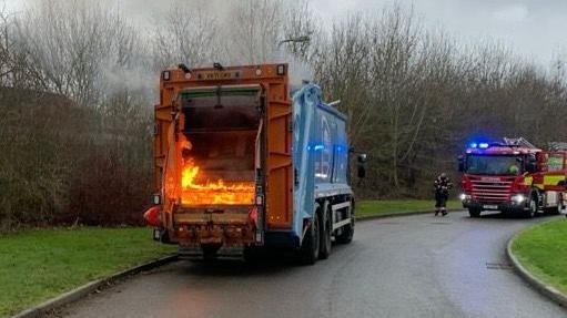 A bin lorry on fire. A fire engine is in the background