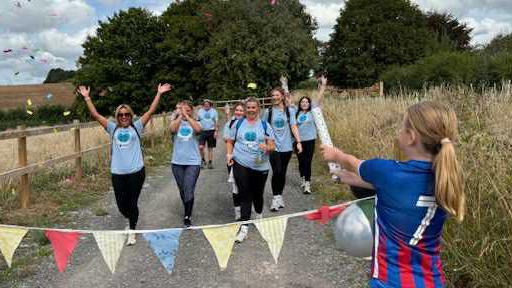 A group of about 15 people wearing dark trousers and light blue T-shirts celebrating as they cross a line of brightly-coloured bunting. In the foreground a child, facing away from the camera, is releasing a mini confetti cannon. The group are walking along a gravel path in the countryside, surrounded by fields and trees.