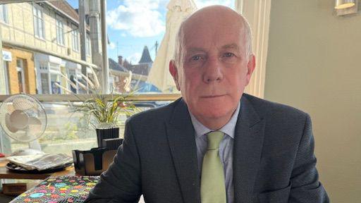 A man looking straight into the camera.  He is wearing a grey suit jacket, pale blue shirt underneath and a pale green tie.  He is sitting next to the bar of a pub. 