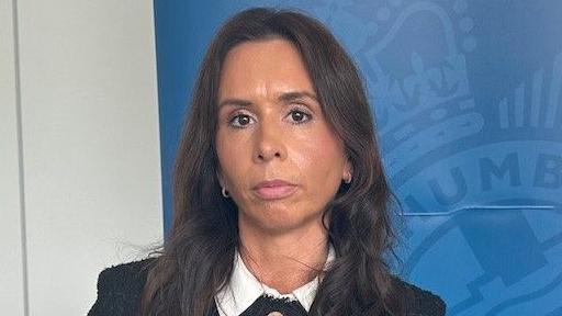 Katie Smith looks at the camera with a serious expression on her face. She has long brown hair and is wearing a dark suit, behind he is a blue banner with the Northumbria Police insignia on it