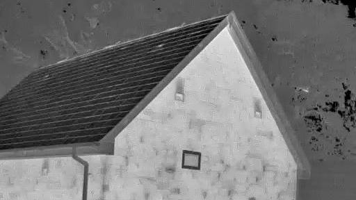 A black and white grainy thermal image of a stone building with three small windows and a slate roof and a speck of bright light to the right which is the bat flying out